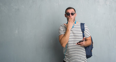 Young caucasian student man over grey grunge wall wearing headphones using smartphone cover mouth with hand shocked with shame for mistake, expression of fear, scared in silence, secret concept