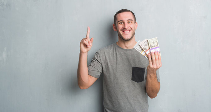 Young caucasian man over grey grunge wall holding a bunch of money surprised with an idea or question pointing finger with happy face, number one