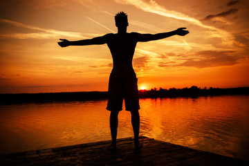 Young man standing  with open arms on the pier by the lake.He joying in summer sunset.