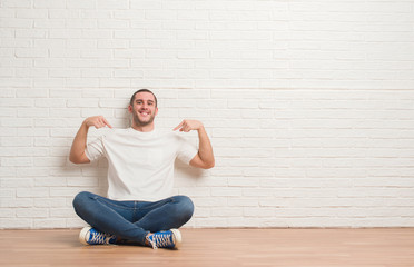 Young caucasian man sitting on the floor over white brick wall looking confident with smile on...