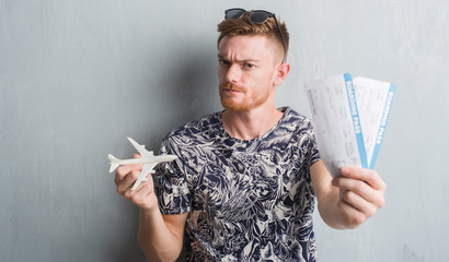 Young redhead man holding aircraf and boarding pass on holidays skeptic and nervous, disapproving...