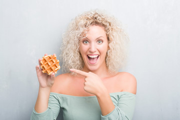 Young blonde woman over grunge grey background eating belgian waffle very happy pointing with hand and finger