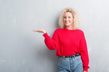 Fototapeta na wymiar Young blonde woman with curly hair over grunge grey background smiling cheerful presenting and pointing with palm of hand looking at the camera.