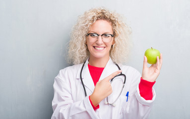 Young blonde dietitian doctor woman over grunge grey wall holding green apple very happy pointing...