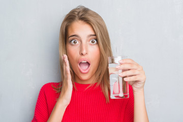Beautiful young woman over grunge grey wall drinking a glass of water scared in shock with a surprise face, afraid and excited with fear expression