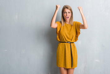 Beautiful young woman standing over grunge grey wall wearing a dress angry and mad raising fist frustrated and furious while shouting with anger. Rage and aggressive concept.