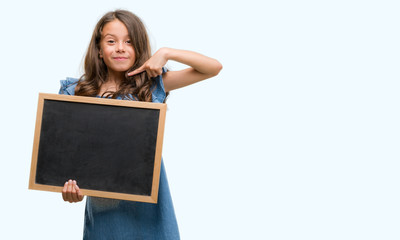 Brunette hispanic girl holding blackboard with surprise face pointing finger to himself