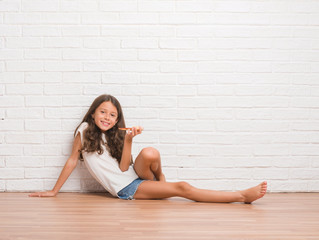 Young hispanic kid sitting on the floor over white brick wall eating pizza slice with a happy face...
