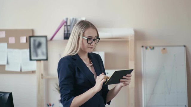 Call center employee at work in the office. Young girl with blonde hair with glasses in the office. The girl looks at the digital tablet, enters data into it, then looks at the camera. The girl stands