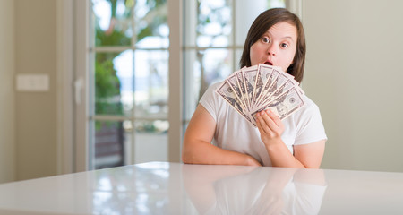 Down syndrome woman at home holding dollars scared in shock with a surprise face, afraid and excited with fear expression