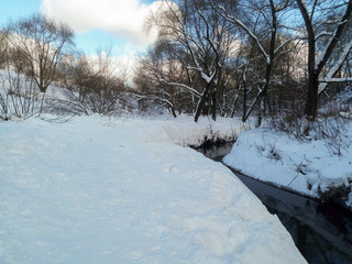 Nonfreezing Ramenka river on a tranquil clear winter day. Moscow. Russia. Fluffy snow adorns leafless trees with festive dresses. Winter nature delights eyes with blinding purity.