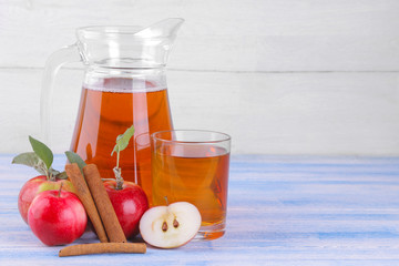 Apple juice in a jug and a glass next to fresh apples and cinnamon sticks on a blue wooden table and on a white wooden background