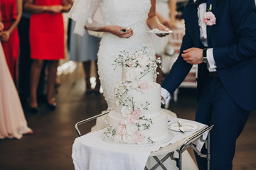 bride and groom tasting stylish white wedding cake with flowers. yummy modern big wedding cake with...