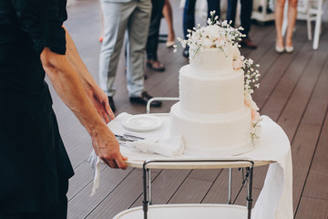 waiter rolling in amazing white wedding cake with flowers. modern white wedding cake with pink and white roses. luxury catering in restaurant. wedding reception.