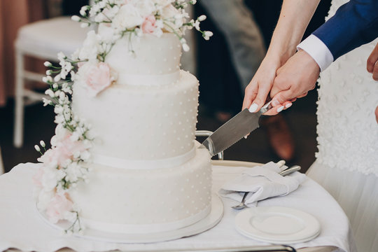 bride and groom holding knife and cutting stylish white wedding cake with flowers. modern big wedding cake with pink and white roses. luxury catering in restaurant. wedding reception