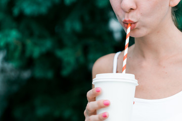 Beautiful young girl stands in a forest with a mug with a red straw. Female hand holding a blank paper cup in a wooded landscape background. Model sucks close up. Mockup for products