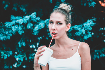 Obraz premium Handsome strong running coach with an ideal fit body holds in his hand a mug with juice and drinks it through a red straw against the background of a green forest. Mockup for products