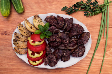 A portion of fried liver with stewed apples, decorated with slices of tomato and sweet pepper, next to a wooden background cucumbers, onions and parsley.
