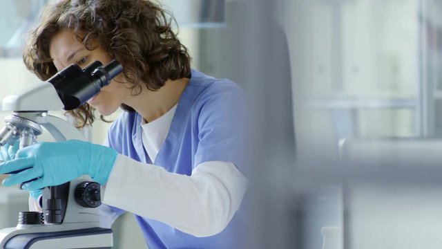 Young female scientist in scrubs and protective gloves looking through microscope while working in laboratory