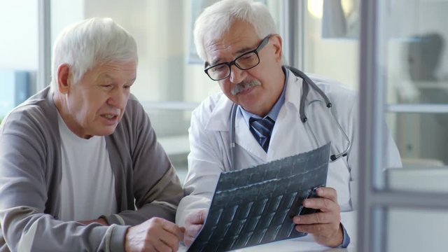 Senior male doctor in lab coat explaining x-ray image to elderly male patient during medical checkup in clinic