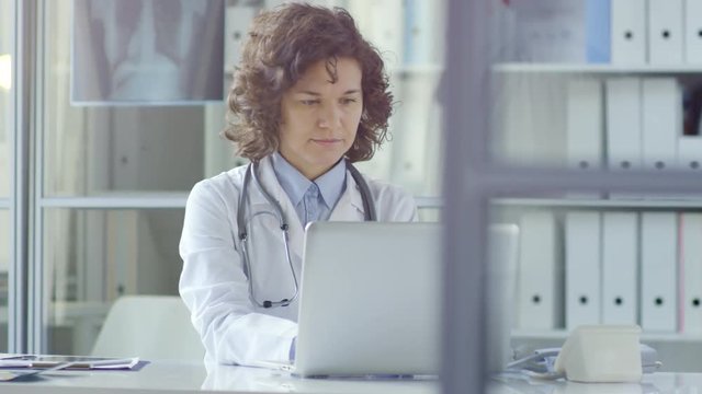 Tracking shot of pretty female doctor in lab coat sitting at desk in modern clinic with glass walls and working on laptop