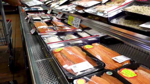 Buying pre-packaged fish at the supermarket. Packs of seafood in open refrigerator at the store. Woman browses chooses and selects various packages to buy.
