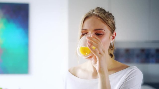 Beautiful girl drinking fresh orange juice from glass. Close up happy woman drinking fruit beverage. Pretty girl in white t-shirt with charming smile standing in kitchen. Healthy and natural nutrition