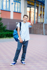 cute, young boy in blue shirt stands  in front of school with blue backpack and thumb up. Education, back to school concept