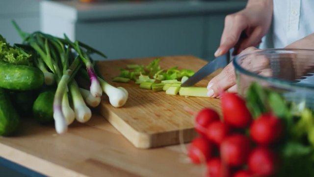 Cooking fresh salad mix with onion, cucumber and radishes. Close up woman hands cutting green onion on wooden board. Fresh vegetables ingredients on kitchen table. Cooking healthy vegetarian food