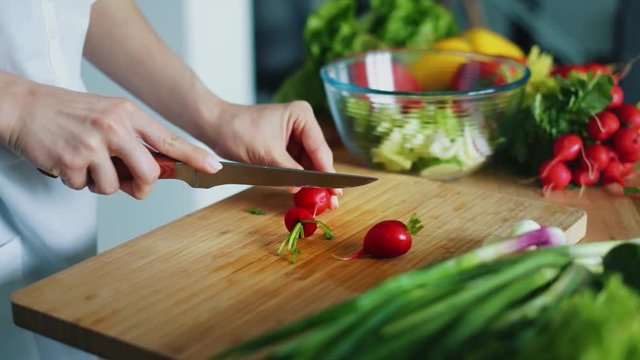 Woman hands with knife cutting radishes for salad. Close up fresh vegetables on kitchen table. Housewife cooking natural and healthy meal on wooden board. Seasonal vegetables ingredients for salad
