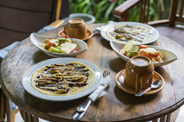 breakfast table setting with banana omelet and fresh mixed fruit salad, with coffee or tea cups in bali homestay
