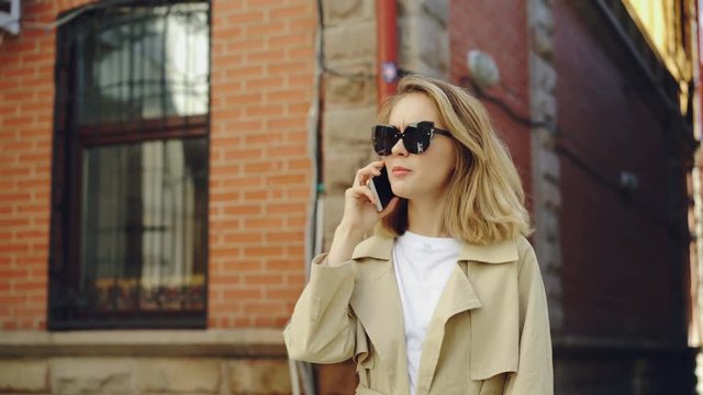 Attractive woman with blond hair is talking on mobile phone walking along street in modern city. Young lady is wearing trendy black sunglasses and summer coat.
