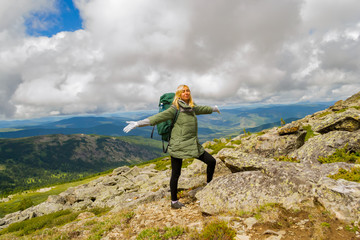 the blond tourist girl in a green jacket and with a backpack standing on a mountain resting and smiling, eyes closed and arms outstretched,against  of mountains, forests and blue sky with white clouds