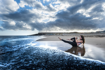 Naklejka premium blue colors epic style image with pretty brunette doing sport and pilates activity at the beach on the shore. scenic wave and clouds sunset on the background. balance lay down position for nice woman