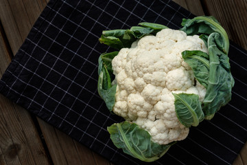 a head of cauliflower with leaves lies on a black in white cage towel on a dark wooden background. view from above