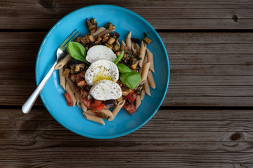 Italian whole-grain pasta penne with eggplants, tomatoes, capers, basil and fresh mozzarella in a turquoise plate on a dark wooden background. view from above