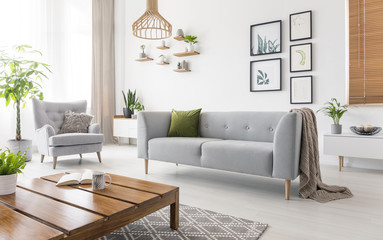 Real photo of grey sofa with green cushion and blanket standing in white living room interior with...