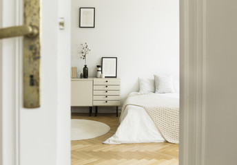 A peek through a door into a monochromatic, white bedroom interior with a bed and cabinets standing...