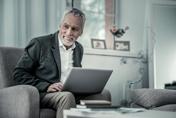 Have idea. Handsome male keeping smile on his face, holding laptop on knees and looking aside