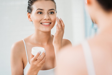 beautiful smiling young woman applying face cream and looking at mirror