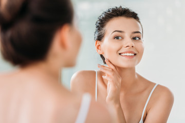 beautiful smiling young woman touching skin and looking at mirror in bathroom - Powered by Adobe