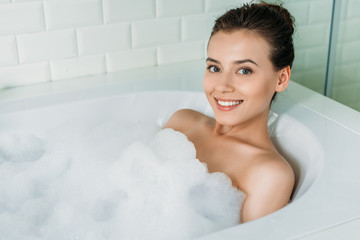 high angle view of beautiful happy girl relaxing in bathtub with foam and smiling at camera