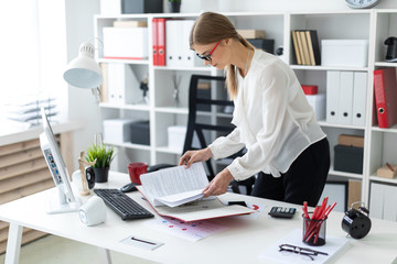Obraz premium A young girl stands bent near the table, holds a pencil in her hand and flips through the folder with the documents.
