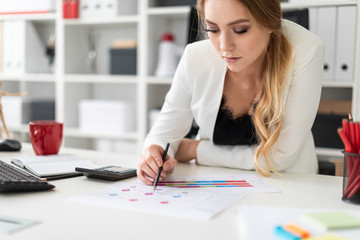 A young girl sits at a computer desk in the office, holds a pencil in her hand and works with charts and diagrams.