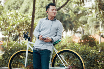 Attractive Asian guy holding smartphone and listening to music while leaning on bicycle on background of park and looking away
