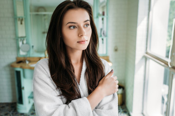 pensive brunette girl in bathrobe looking at window in bathroom
