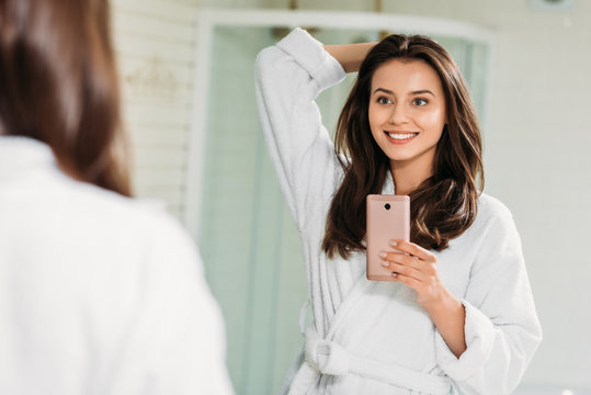 beautiful smiling young woman in bathrobe taking selfie with smartphone in bathroom