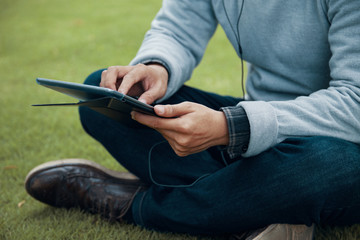 Anonymous guy in casual outfit sitting on green lawn and using modern tablet