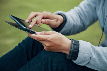 Side view of unrecognizable man in casual outfit using modern tablet while sitting on ground in park