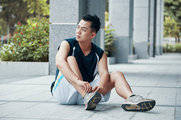 Attractive Asian guy in sportswear tying laces on sneakers and looking away while sitting on ground...